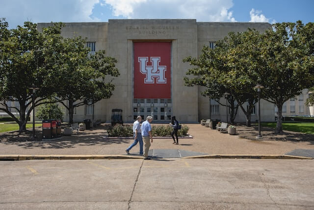 Individuals can be observed walking outside a building situated at the University of Houston.